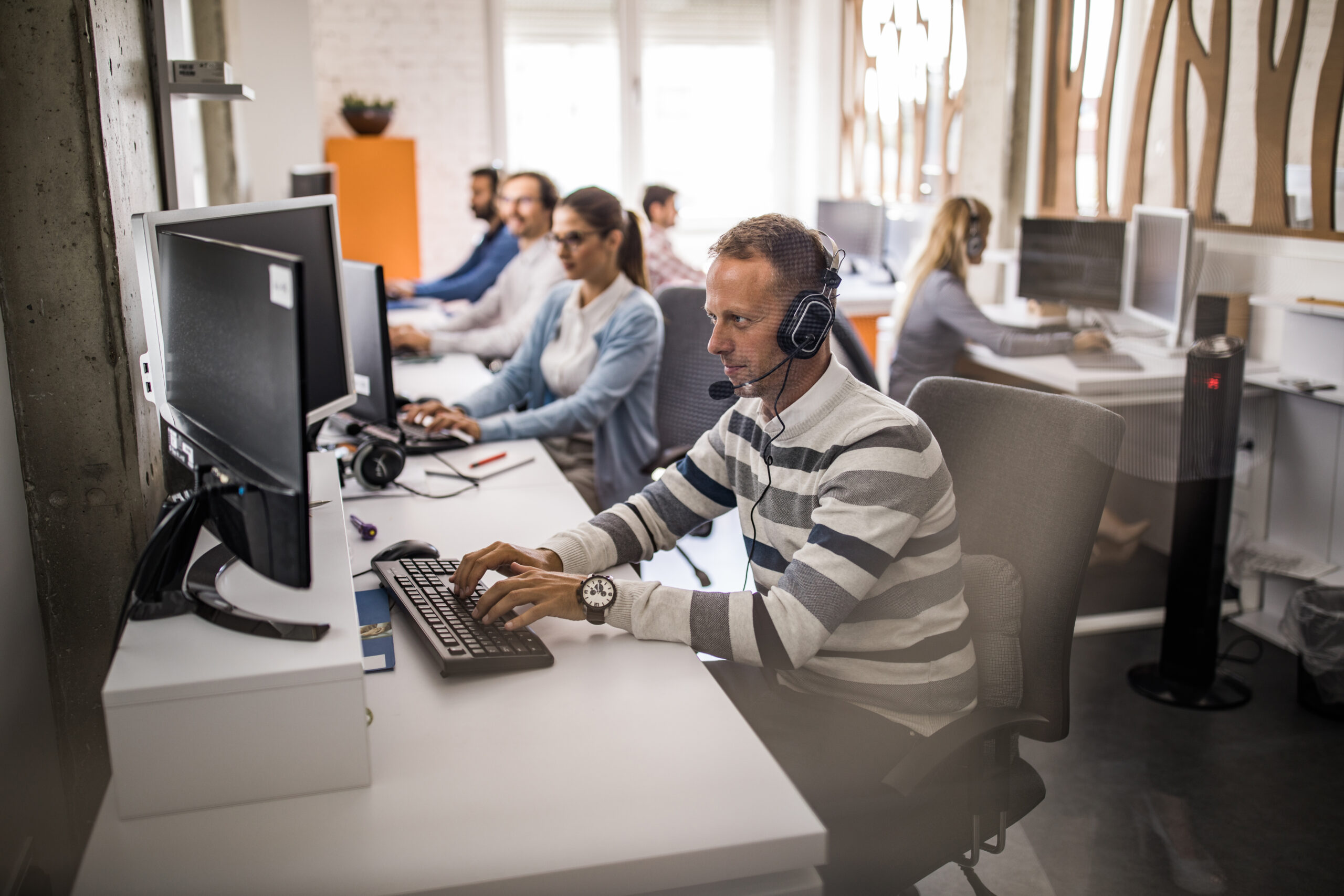 Young man working on desktop PC at call center with his colleagues. The view is through glass.